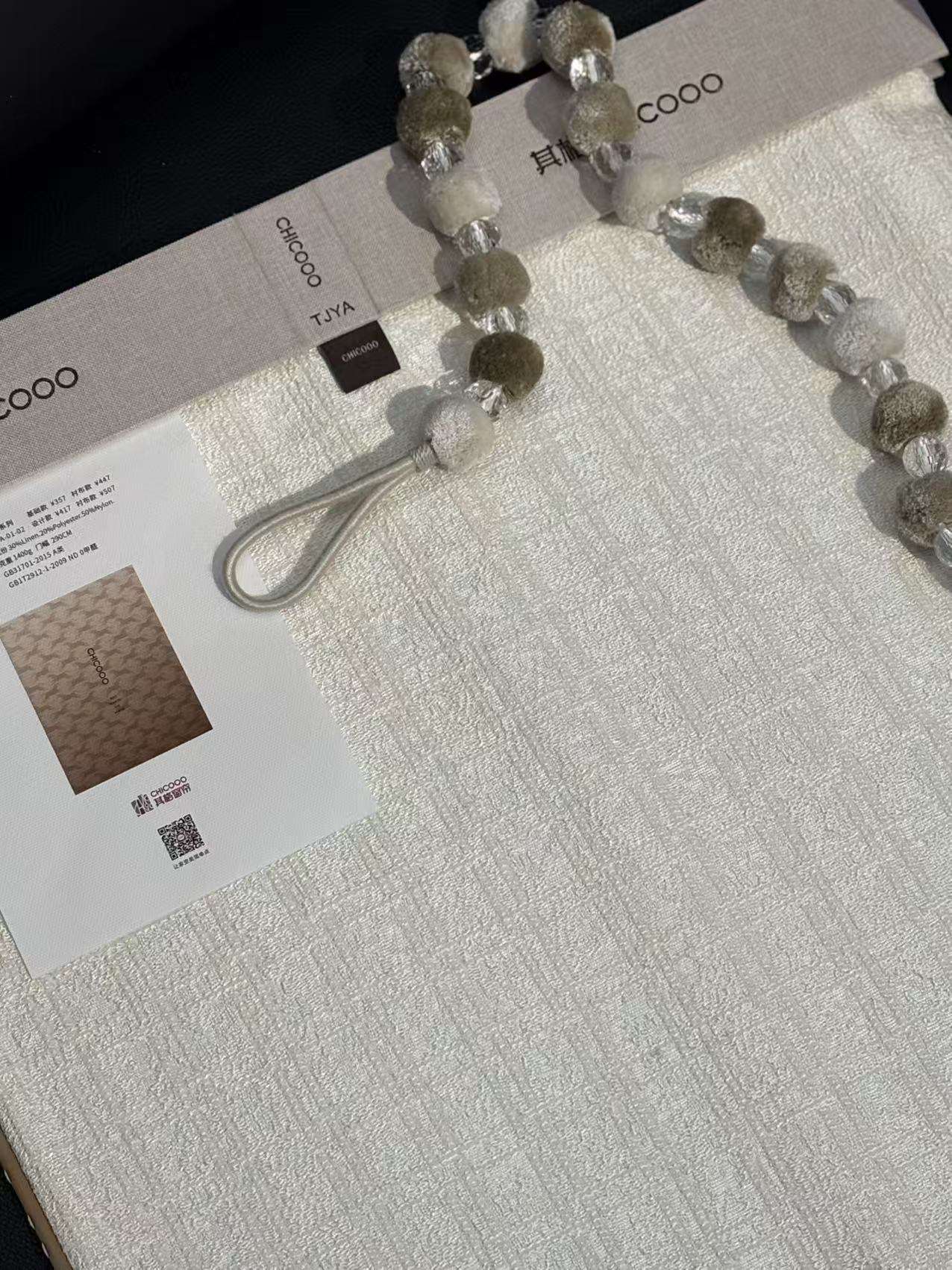 这是一张展示CHICOOO其趣窗帘面料样册的特写图片。画面主体为一块白色肌理感窗帘布,表面呈现细腻的竖条纹雪尼尔质感,搭配一串由灰绿色毛绒球和透明水晶珠交替串成