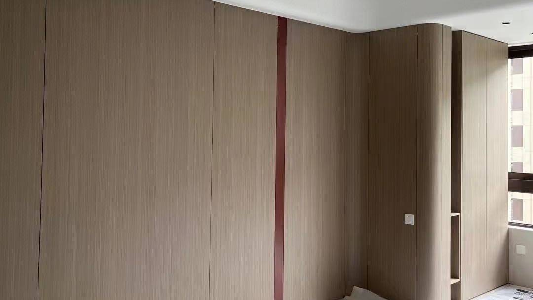 室内实景展示浅木色木饰面护墙板与定制柜体。采用细腻竖向木纹，搭配柔和的圆弧转角设计，消除空间锐利感。中间嵌入红褐色装饰线条，右侧设有开放式层板与白色开关面板。整